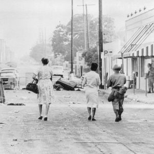 LOS ANGELES, CA--August 15, 1965--A rifle-carrying National Guardsman escorts two women through the debris-littered streets of Watts where rifle fire from snipers continued to harass Negroes and whites alike in Los Angeles' riot zone. The women were bound for Sunday church services. (AP Wirephoto.)